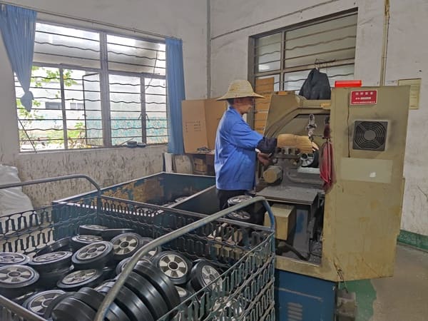 A factory worker wearing a straw hat operating a machine to process rubber wheels, with storage bins filled with finished rubber wheels nearby, in a workshop .