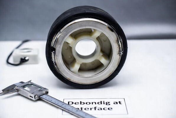 Rubber wheel debonding at interface, cross-section with steel hub, caliper measurement on white bench, bond failure inspection.